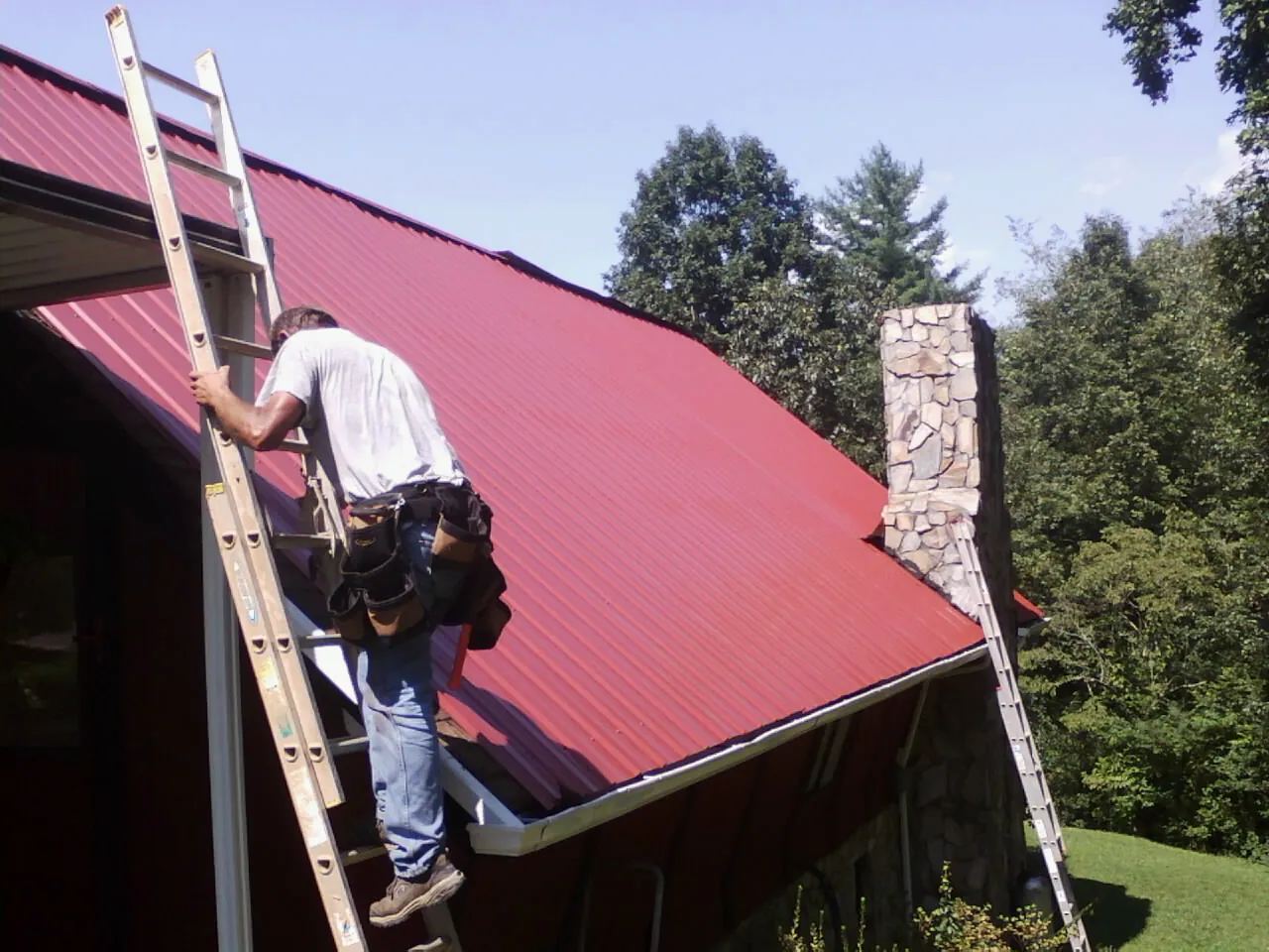 Roofer on a ladder beside a red metal roof and stone chimney — Soffit & Fascia Repair by Roofing Brothers in Warden
